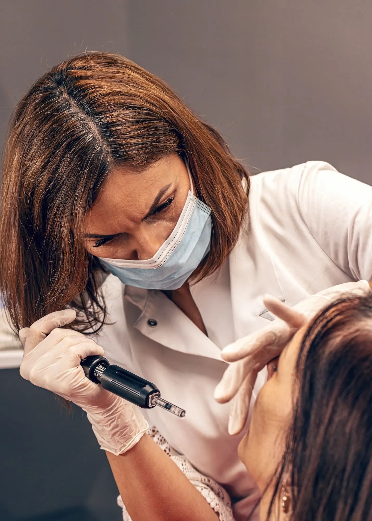 Behandlerin mit Mundschutz behandelt die Lippen einer Kundin mit einem Stiftgerät