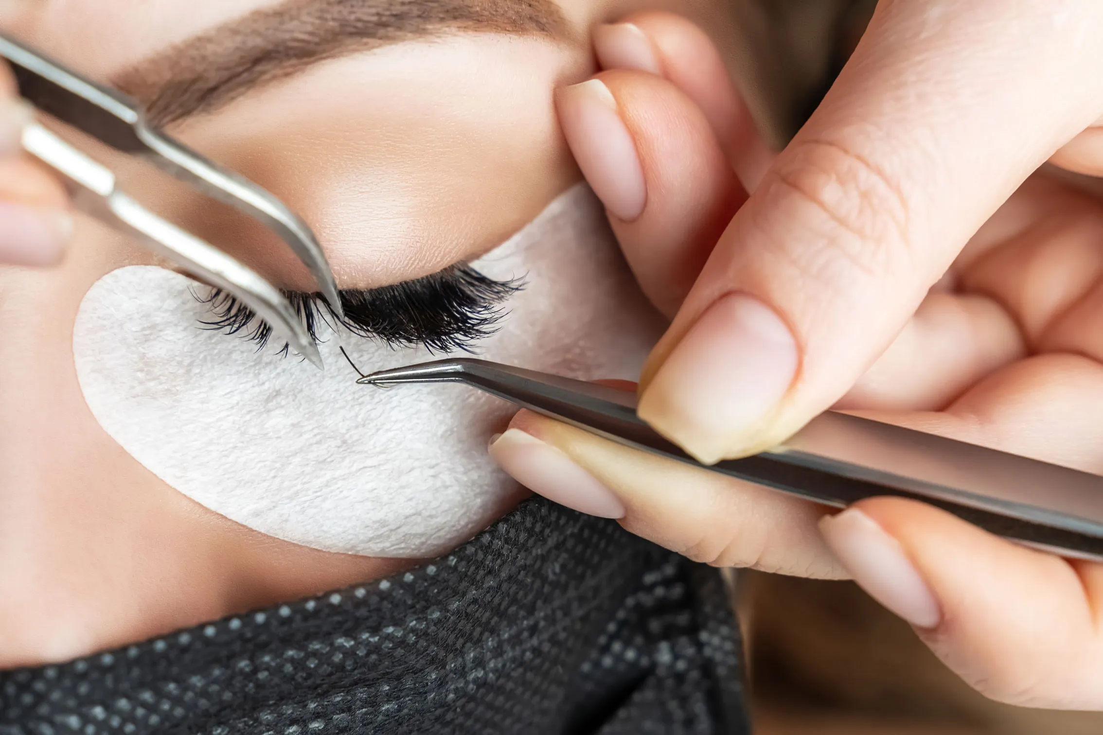 Einzelne Wimper wird mit zwei Pinzetten am geschlossenen Auge angesetzt
